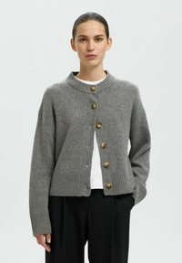 Grauer gestrickter Cardigan mit hohem Kragen, gerippten Bündchen und sechs goldfarbenen Knöpfen an der Vorderseite. Über einem weißen Hemd getragen.