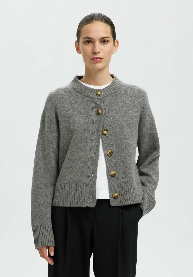 Grauer gestrickter Cardigan mit hohem Kragen, gerippten Bündchen und sechs goldfarbenen Knöpfen an der Vorderseite. Über einem weißen Hemd getragen.