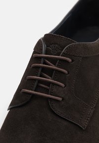 Zapato de ante marrón con un sistema de cordones cerrado, costuras distintivas y un logo sutil grabado en la parte superior cerca de la zona de los cordones.
