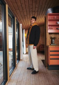 Black bomber jacket over a mustard hoodie, light beige trousers, and black shoes, standing in a wooden interior with orange cabinetry.