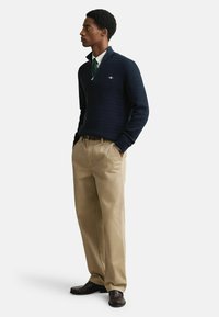 Marineblauwe gebreide trui met een kwart-rits kraag, beige relaxte pasvorm broek en donkere schoenen, met een subtiel logo op de trui.