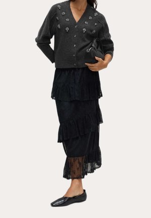 Femme portant un cardigan gris foncé orné de perles, une jupe noire à volants en dentelle, des ballerines noires, et tenant un petit sac à main noir à hauteur de taille.