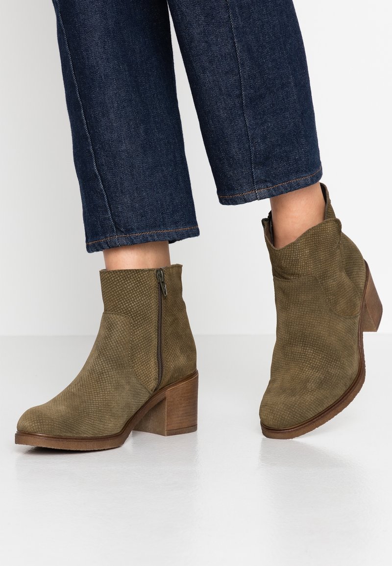 Bottes en daim vert olive arrivant à la cheville, avec fermeture éclair latérale et talon bloc, portées avec un jean court bleu foncé.
