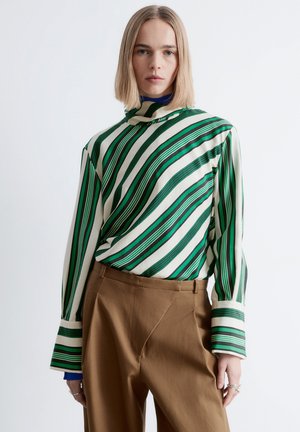 Kvinde med glat blondt hår iført en grøn og hvid diagonalt striped bluse med brede manchetter og høj hals, kombineret med brune bukser.