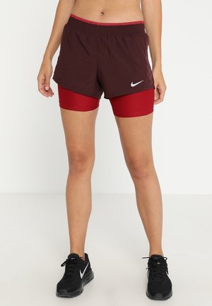 Vrouw die gelaagde donkerrode sportshorts draagt met daaronder rode compressieshorts, en zwarte Nike hardloopschoenen.