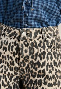 Jean en denim à imprimé léopard avec une base beige clair et des taches noires, doté d'une fermeture par bouton argenté et de détails cousus.