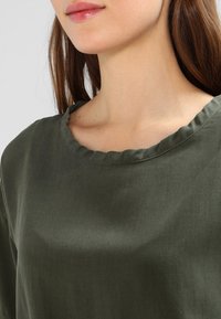 Dark green blouse with a rounded neckline, featuring a scalloped edge and smooth texture, partially visible worn by a model.