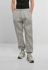 Gray sweatpants with elastic waistband, tapered cuffs, side pockets, and embroidered branding on the left hip. Worn with silver sneakers.