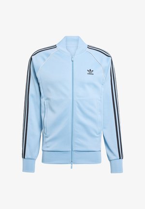 Giacca Adidas azzurro chiaro con zip frontale, polsini a costine, tre strisce nere sulle maniche e un piccolo logo nero sul lato sinistro del petto.