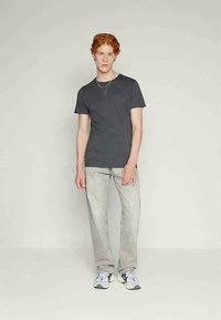 Jonge man met rood haar staat gekleed in een donkergrijze t-shirt, lichtgrijze jeans, witte sneakers en gelaagde zilveren kettingen tegen een effen achtergrond.