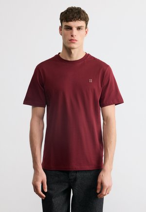 Tânăr în picioare, purtând un tricou simplu bordo cu mâneci scurte și un mic logo „II” pe piept, și blugi închiși la culoare.