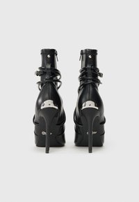 Pár čiernych vysokých členkových topánok na podpätku s zadnými zipsami, obalenými popruhmi a striebornými kovovými akcentmi na jednoduchom bielom pozadí.