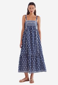 Robe maxi bleue avec des motifs de feuilles blanches, bretelles réglables et bas à volants. Fabriquée en tissu léger avec des détails soignés.