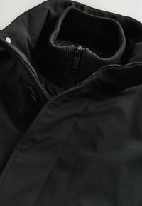 Chaqueta negra con cuello alto acanalado, fabricada en un tejido suave, con cremallera oculta y botones a presión, de diseño minimalista y sin patrones visibles.
