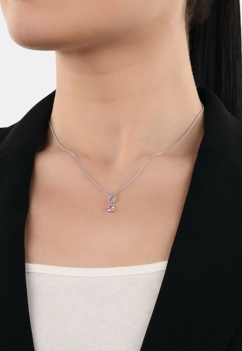 Silverfärgad kedjepyssel med en rosa hängsmycke, prytt med en rundslipad ädelsten. Enkel design mot en svart kavaj och vit topp.