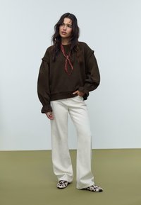 Felpa marrone oversize con maniche a puff, abbinata a pantaloni larghi color crema e scarpe slip-on a pois. Foulard come dettaglio.