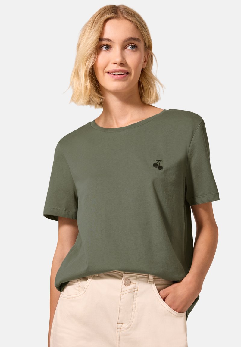 Femme blonde portant un t-shirt vert olive avec un petit logo cerise noir et un pantalon beige clair, main dans la poche, souriant légèrement.