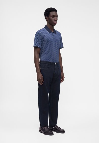 Jeune homme debout portant un polo bleu à motifs, un jean foncé et des mocassins en cuir noir sur un fond blanc uni.