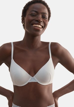 Femme souriante portant un soutien-gorge blanc et une lingerie en dentelle, les mains sur les hanches, sur un fond clair uni.