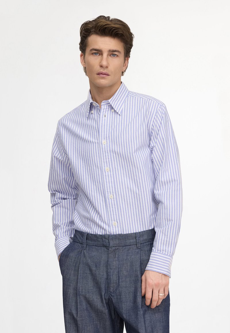 Jeune homme aux cheveux châtain clair portant une chemise boutonnée rayée bleue et blanche et un pantalon gris foncé, une main dans la poche.