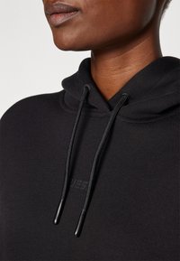 Felpa nera con cappuccio, dotata di tasca a marsupio frontale, cordini con punte in plastica e un logo goffrato discreto sul petto. Tessuto dalla texture liscia.