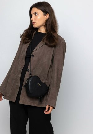 Schwarze Leder-Handtasche in Herzform mit strukturiertem Oberflächen und goldenen Beschlägen, quer über die Schulter getragen. Modell in braunem Wildlederblazer und schwarzem Oberteil.
