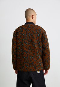 Brun, fluffy jakke med grøn- og sort leopardmønster, der har en ribbet sort krave og et lille hvidt logo på ryggen.