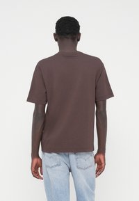 Rjava majica s kratkimi rokavi iz mehke bombažne tkanine, z klasičnim okroglim ovratnikom in sproščenim krojem, kombinirana s svetlo modrimi jeans hlačami.