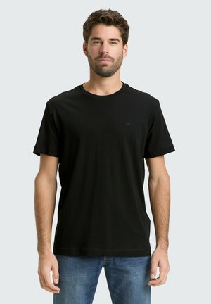 Man standing facing forward wearing a plain black t-shirt and blue jeans against a light gray background.