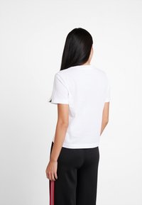 A woman with long black hair wears a white t-shirt and black pants with red stripes, seen from the back against a plain background.