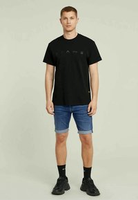 Homme debout, portant un tee-shirt noir, un short en jean retroussé, des chaussettes noires et des sneakers noires, contre un fond clair.