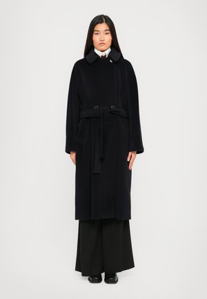 Woman with long dark hair wearing a black belted coat over wide-leg black pants and black shoes, standing against a plain white background.