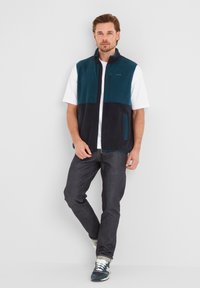 Man wearing teal and navy fleece vest over white short-sleeve shirt with dark jeans and blue sneakers, standing against plain light gray background.