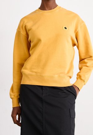 Osoba ubrana w żółtą bluzę Carhartt crewneck i czarną spódnicę z boczną kieszenią, stojąca z jedną ręką w kieszeni.