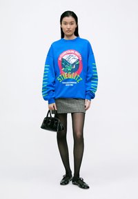 Donna che indossa un felpa blu brillante con grafica di montagna, gonna in pied-de-poule bianco e nero, collant neri, ballerine nere, e tiene una borsa nera.