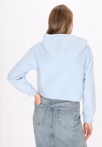 Felpa blu chiaro tagliata, con cappuccio e polsini elastici, indossata sopra una gonna di jeans con tasche posteriori, mostrando una morbida texture.