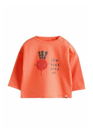 Koralinė medvilninė marškinėlių su ilgomis rankovėmis, puošta raudono burokėlio grafika su juodais lapais ir užrašu „the beet goes on“. Kortelės užsegamos prie peties.