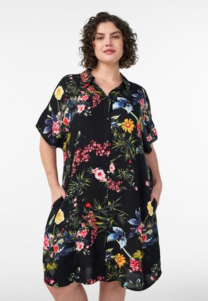 Donna con capelli corti e ricci che indossa un vestito nero con maniche corte e bottoni, decorato con motivi floreali colorati, con le mani nelle tasche.