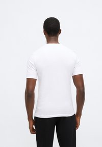 Balti medvilniniai trumpomis rankovėmis marškinėliai su apvaliomis kaklo apykaklėmis ir lygiu paviršiumi. Dėvimi su juodomis tekstūruotomis kelnėmis, matomi iš nugaros.