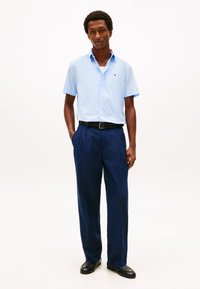 Mann iført lys blå kortermet skjorte med knapper, mørkeblå bukser, svart belte og svarte sko, stående med hendene i lommene mot hvit bakgrunn.