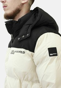 Giacca a vento nera e crema, con colletto alto, bottoni a pressione e patch con logo sulla manica. Materiale testurizzato con design trapuntato.