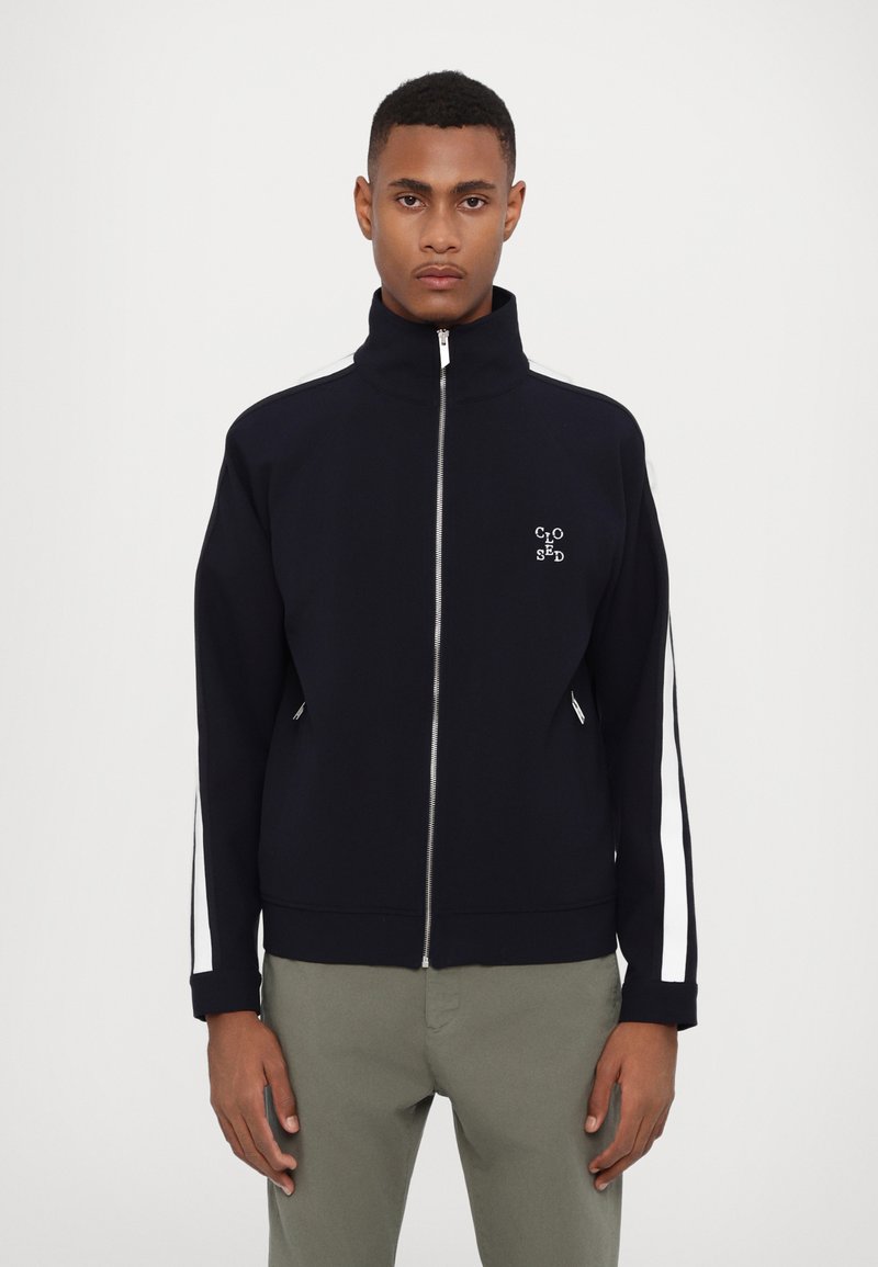 Veste zippée marine avec col haut, poches latérales zippées, rayures blanches le long des manches et un petit logo sur la poitrine. Texture de tissu lisse.