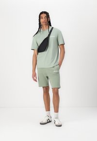 T-shirt in cotone verde chiaro e pantaloni corti abbinati, con una borsa a tracolla nera. Calze bianche e scarpe da ginnastica grigie con accenti neri. Design semplice.