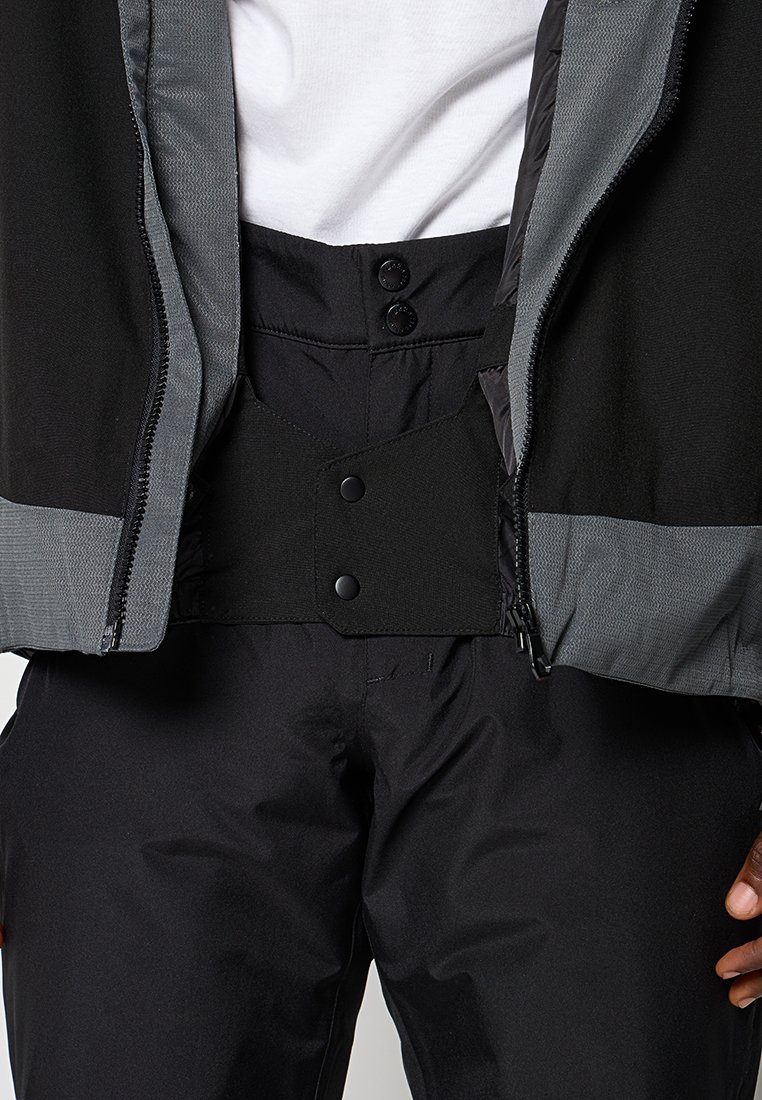 Pantalones de nieve negros de cintura alta con cierre de botones a presión ajustable, combinados con una chaqueta negra y gris con cremallera y una camiseta blanca debajo.