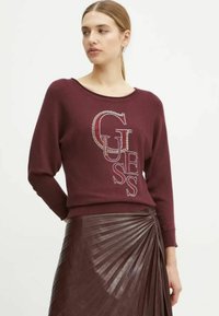 Felpa bordeaux con maniche lunghe, presenta lettere testurizzate "GUESS" impreziosite da strass, abbinata a una gonna in pelle plissettata.
