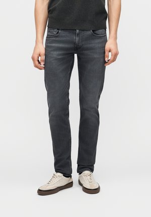 Άνδρας που φοράει σκουρόγριμο τζιν στενής γραμμής και λευκά κα casual sneakers, στέκεται μπροστά σε ένα απλό φωτεινό φόντο.