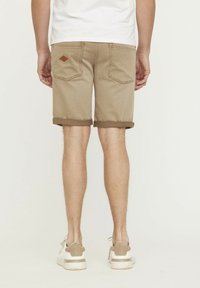 Beige Shorts mit umgeschlagenem Saum und Gesäßtaschen, mit einem Rautenmuster-Detail. Getragen mit hellen Sneakers vor einem neutralen Hintergrund.