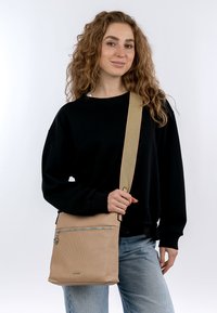 Borsa a tracolla in pelle beige con finitura testurizzata, zip argentata, tasca frontale e manico beige regolabile. Modello che tiene la borsa in primo piano su uno sfondo bianco.