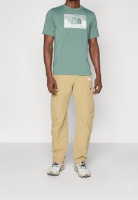 Zielona bawełniana koszulka z grafiką North Face, w połączeniu z beżowymi lekkimi spodniami z kieszenią na zamek oraz szarymi butami do wędrówek.