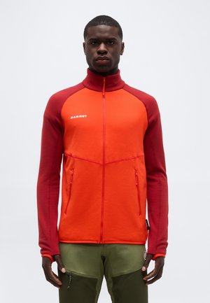 Vyras dėvi raudonos ir oranžinės spalvų „Mammut“ su užtrauktuku striukę su užtrauktukais kišenėse ir alyvuogių žalios spalvos kelnes, stovintis priešais paprastą foną.